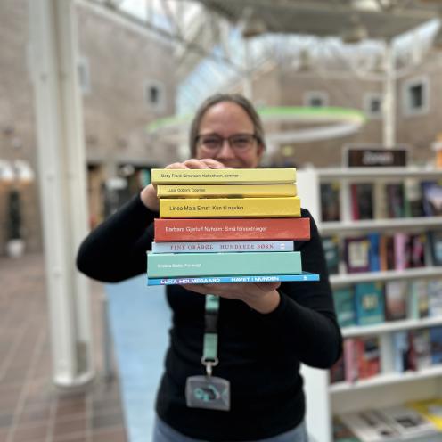 Tine står blandt fyldte bogreoler på Hovedbiblioteket. Hun holder syv af de omtalte bøger i denne artikel frem for sig i en stak med bogryggene frem. Der er fokus på bogryggene, mens både Tine og resten af baggrunden, som viser biblioteksrummet, fremstår let sløret. Man kan ane, at Tine smiler.