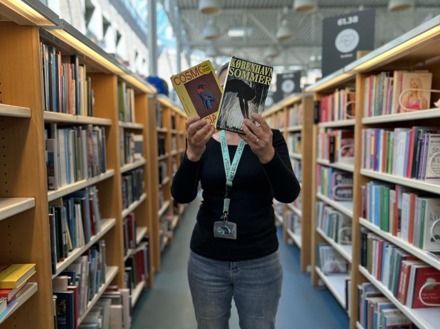 Tine står blandt fyldte bogreoler på Hovedbiblioteket. Hun holder to af de omtalte bøger i denne artikel frem for sig, så der er fuld fokus på bogforsiderne, mens både Tine og baggrunden, som viser biblioteksrummet, fremstår let sløret.